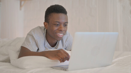 Young African Man Doing Video Chat on Laptop while Lying on Stomach in Bed