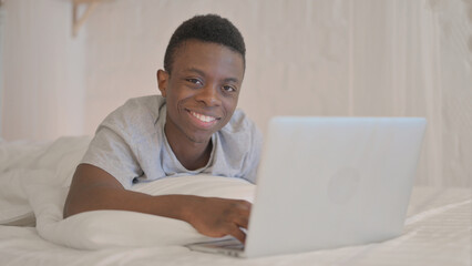 Smiling Young African Man Looking in Camera while using Laptop Lying on Stomach