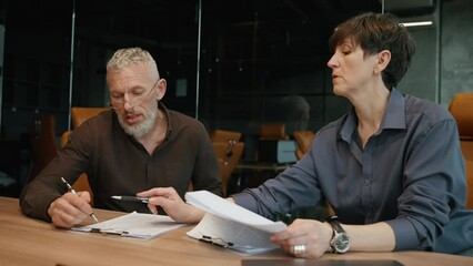 Serious old man and woman colleagues discussing work and signing documents in office