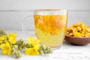 Herbal medicinal tea drink made of Verbascum thapsus, the great mullein, greater mullein or common mullein. Yellow dried flower petals. Glass cup with tea on white background and dried petals in bowl.