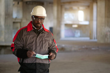 African american builder stands at construction site and holds medical mask