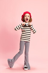 Portrait of happy, excited little girl, cild in striped sweater and red beret posing with vintage photo camra agaisnt pink studio background. Concept of childhood, emotions, fun, fashion, lifestyle