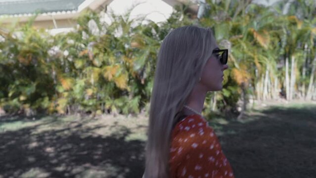Blonde Woman in Red Dress and Sunglasses Walking Along Greenery with Palm Trees on Sunny Day