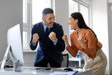 Excited colleagues gesturing YES, celebrating business success in front of computer in office, copy...