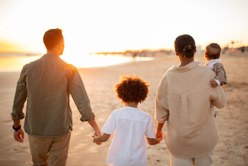 Back view of family walking on the beach with two kids, holding hands, enjoying sunset on seaside