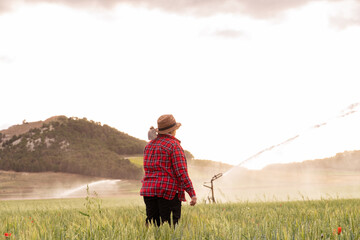 grandmother dressed in straw hat and red plaid shirt holds her hoe with her shoulder and watches her crops being irrigated . Backgrounds of people cultivating in the field.