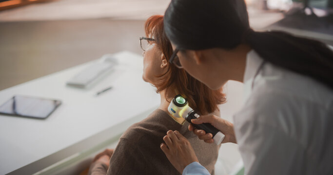 Young Asian Dermatologist is Using a Dermatoscope to Identify Worrying Cancerogenic Tissues on the Skin of a Senior Female During a Health Check Visit to a Clinic. Female Doctor Working in Hospital