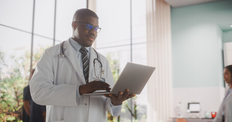 Young African Male Doctor Wearing White Coat, Using a Laptop Computer while Standing in Hospital Hallway. Handsome Black Medical Health Care Professional Working with Patient Test Results
