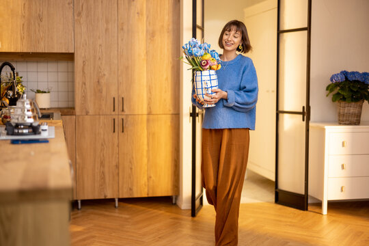 Young woman carries flowers in vase, decorating kitchen interior with fresh flowers or having some household chores