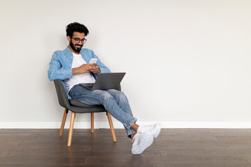 Online Communication. Smiling Indian Guy Using Laptop And Messaging On Smartphone Indoors
