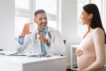 Fototapeta na wymiar Vitamin prescription. Smiling male doctor demonstrating prescribing medications to pregnant woman after check up