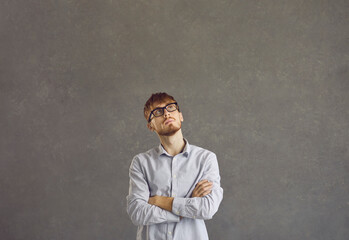 Portrait of a serious caucasian young man with a pensive expression on a gray background. Man in a shirt and glasses stands with folded arms and looks up thinking about what decision to make.