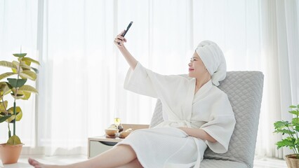 Beautiful young Asian woman in bathrobe and towel turban and using smartphone for taking a selfie...