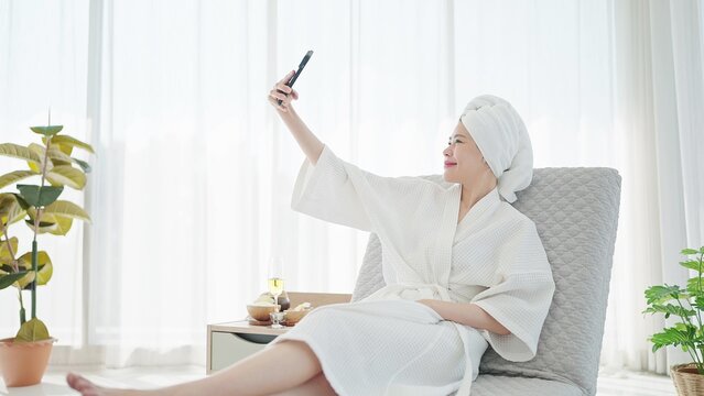 Beautiful young Asian woman in bathrobe and towel turban and using smartphone for taking a selfie or video call while resting on sofa bed in luxury bedroom at home on weekend