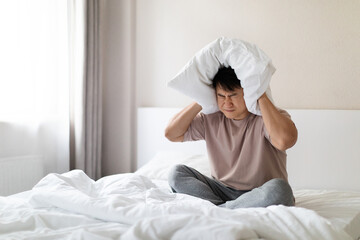 Sleepless man sitting in bed, covering ears with pillow