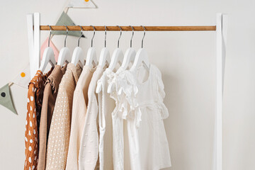 Fashion dresses and jumpers in white, beige and brown colors on hangers in wardrobe. Wooden Clothing Rack with children's outfits. Home kids wardrobe. Set of kids clothes and accessories.