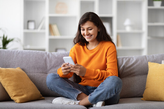 Happy Eastern Woman Sitting On Couch At Home, Using Phone