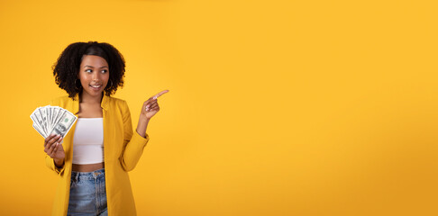 Smiling black curly lady holding lot of money and pointing finger aside at free space on yellow background, panorama