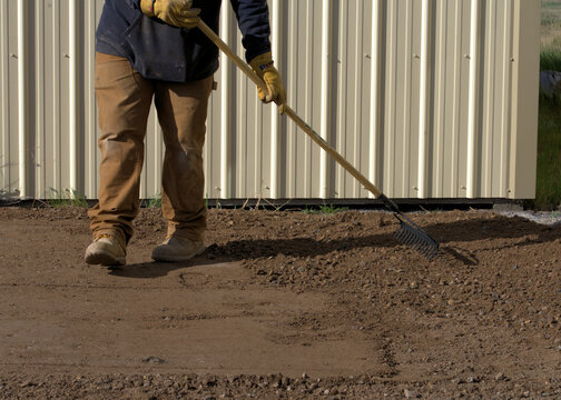 Construction worker walks as he smooths loose earth with a  lawn rake.