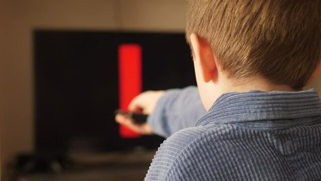 the boy is watching the netflix channel on the tv holding the tv remote, the screensaver of the netflix company on the tv screen. 