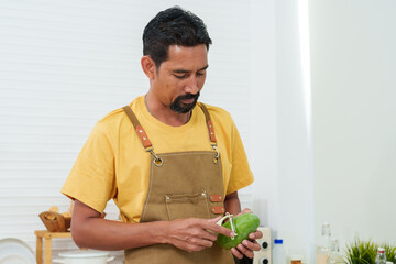 Asian man with mustaches standing in the kitchen in front of the table preparing breakfast The table was full of vegetables, eggs and bread. man wearing an apron stood happily peeling a mango.