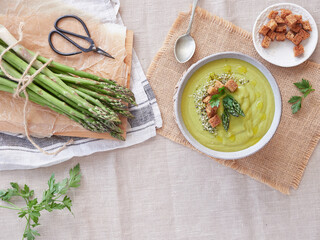 healthy creamy green asparagus soup with pieces of bread croutons next to a bunch of freshly picked green wild asparagus, parsley, spoon and scissors Top view.