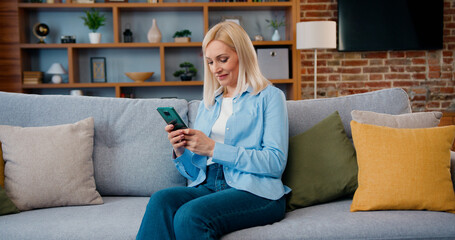 Obraz premium Smiling middle aged lady customer texting mobile sms message playing mobile games using social media apps at home. Millennial woman spending time at home with cell gadget technology.