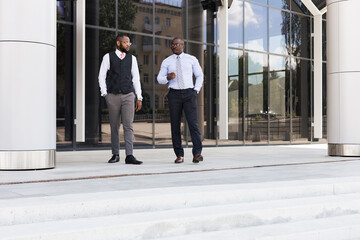 Portrait of two dark-skinned businessmen walking and talking in front of a modern building exterior. Friendly meeting outdoors