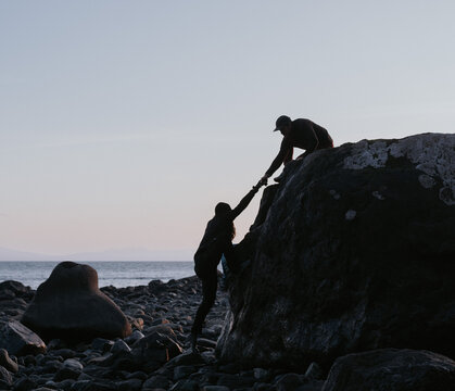 Man helps woman climb a boulder on the beach