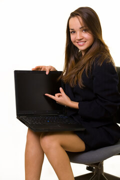 Attractive young brunette business woman sitting on a chair wearing black business suit while pointing to the screen of a laptop computer with friendly expression