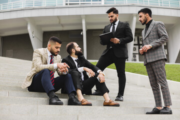 Business people outdoor meeting. A company of male businessmen in suits are sitting on the steps of the stairs. Working break. Discussion and conversation.