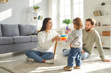 Young happy smiling married couple sitting on the floor with their cute liitle daughter playing an educational pyramid game in the living room at home together. Family and people concept.