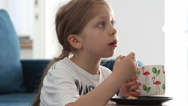 A small blonde girl sits by the sofa with food and watches TV without blinking. Eat food while watching TV. It's okay to look at the screen. The child is watching TV.