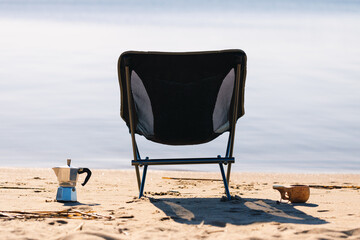 Camping chair, coffee maker and mug in the morning on the seashore close-up background is blurred.