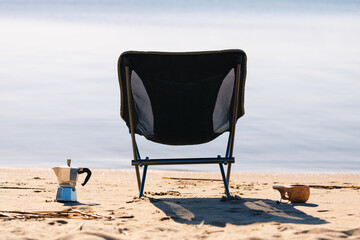 Fototapeta na wymiar Camping chair, coffee maker and mug in the morning on the seashore close-up background is blurred.