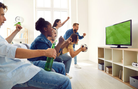 Diverse friends spending weekend together and watching football match at home. Happy excited mixed race people sitting on sofa, looking at TV screen and screaming. Watching soccer concept. Side view