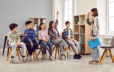 Group of school children sitting in a row in the class room and answering questions of a peer student girl who acts as a teacher. Back to school, education, discussion concept - obrazy, fototapety, plakaty