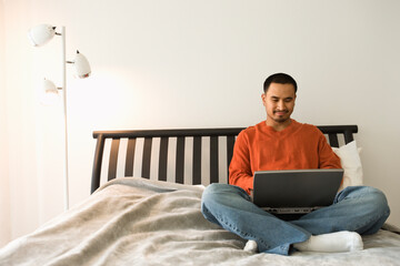 Young Hispanic man sitting cross-legged in bed and using a laptop computer. Horizontal shot.