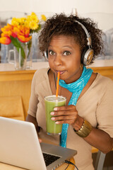 A young woman sitting in a cafe and drinking a frozen beverage. She has a laptop open on the table in front of her and she is wearing headphones draped around her neck. Vertical shot.