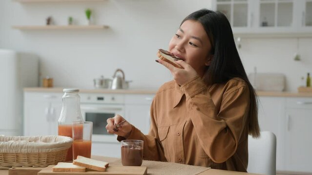 Asian japanese korean chinese woman in kitchen cooking breakfast apply chocolate paste on bread toast ethnic happy girl eat sweet high calorie sandwich enjoy eating make food order delivery at home