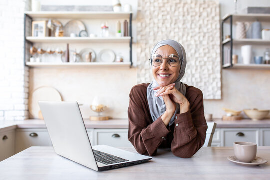 Portrait of a young woman in a hijab sitting dreamily and thoughtfully in the kitchen at home near a laptop. He studies online, works remotely, rests.