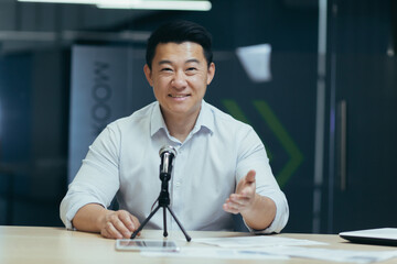 Asian smiling young man sitting in office at desk in front of camera, talking on microphone via...