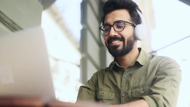 Attractive smiling young indian businessman freelancer with headphones working on laptop and listen to music at coffee shop Relaxed man have distance remote freelance work on computer outdoors 