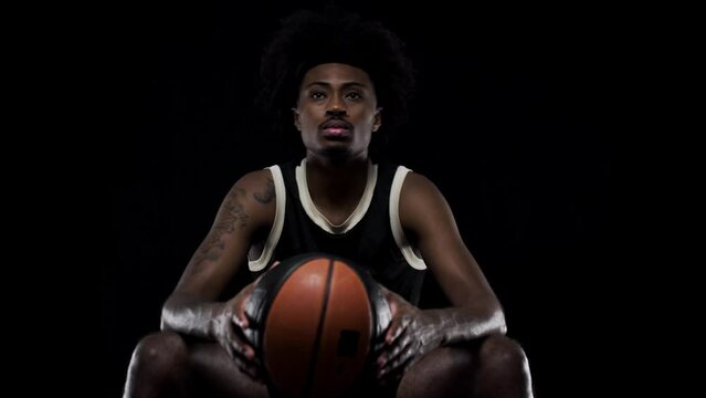 Basketball player sitting on a bench holding a ball. Concentrated african american sports man against black background