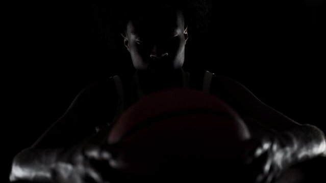 Sidelit basketball player silhouette sitting on a bench holding a ball. Concentrated african american sports man sidelit against black background.