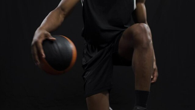 Basketball player bouncing a ball. African american sports man practicing against black background..