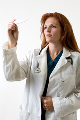 Female attractive red hair doctor wearing white lab coat holding and looking at a  thermometer with a concerned facial expression standing on white background