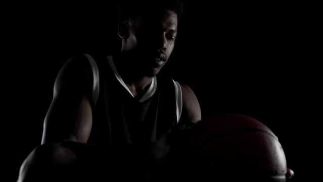 Basketball player sitting on a bench taking deep breath in. Concentrated african american sports man against black background.