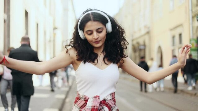 Beautiful young curly indian girl walking down the street and dancing enjoying great day outdoors Smiling female listening to music in headphones at the city street