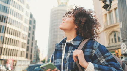 Happy girl using app on smartphone while traveling. Young smiling woman explores the streets, looks up